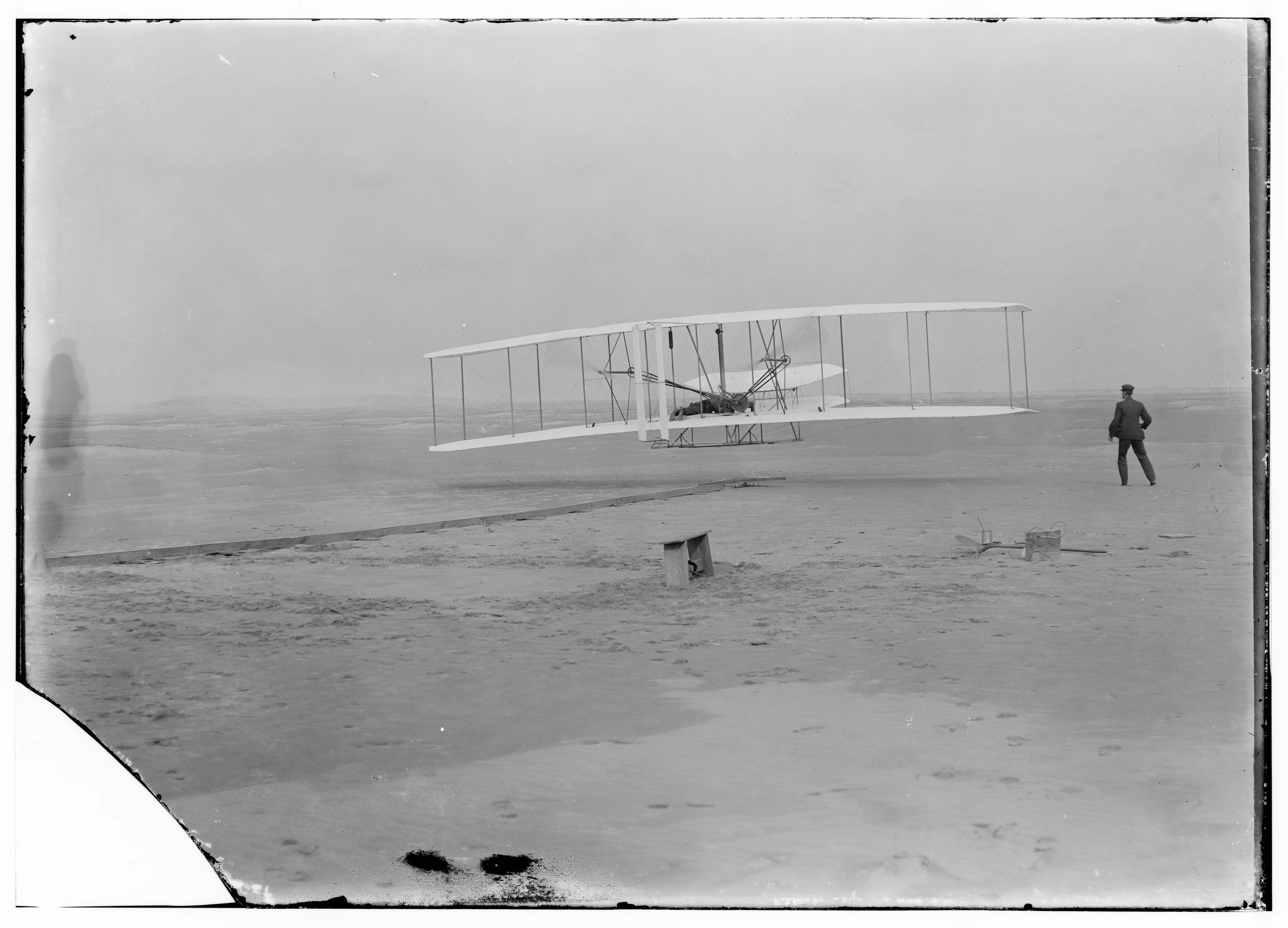 An early aircraft just above the ground, with one person at the controls and another alongside.