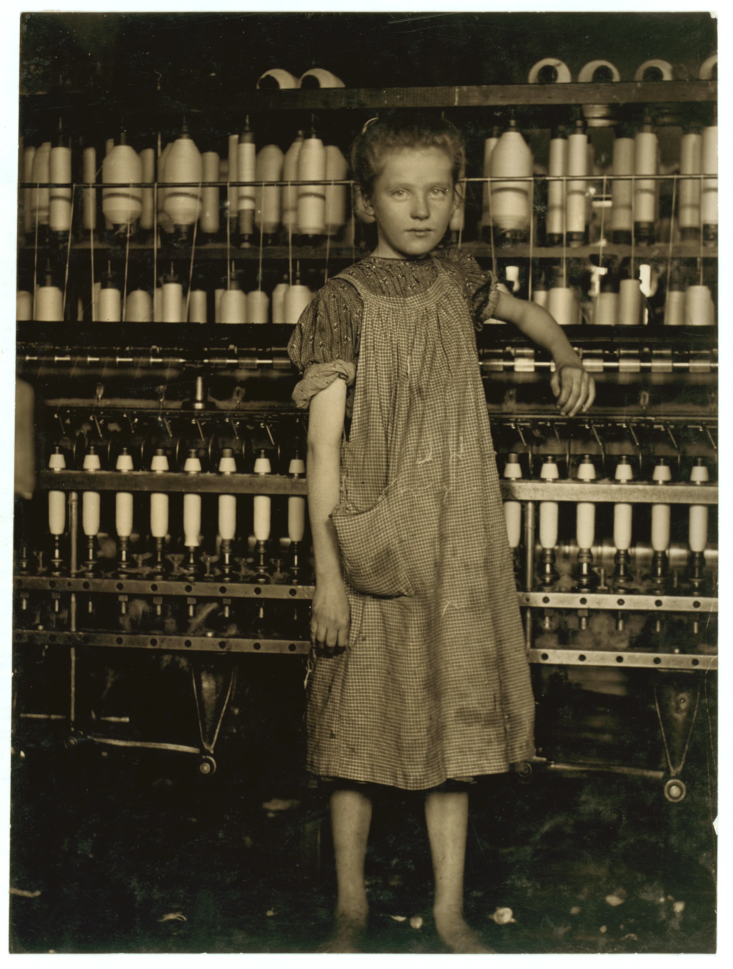 A girl stands beside cotton-mill machinery that extends behind her.