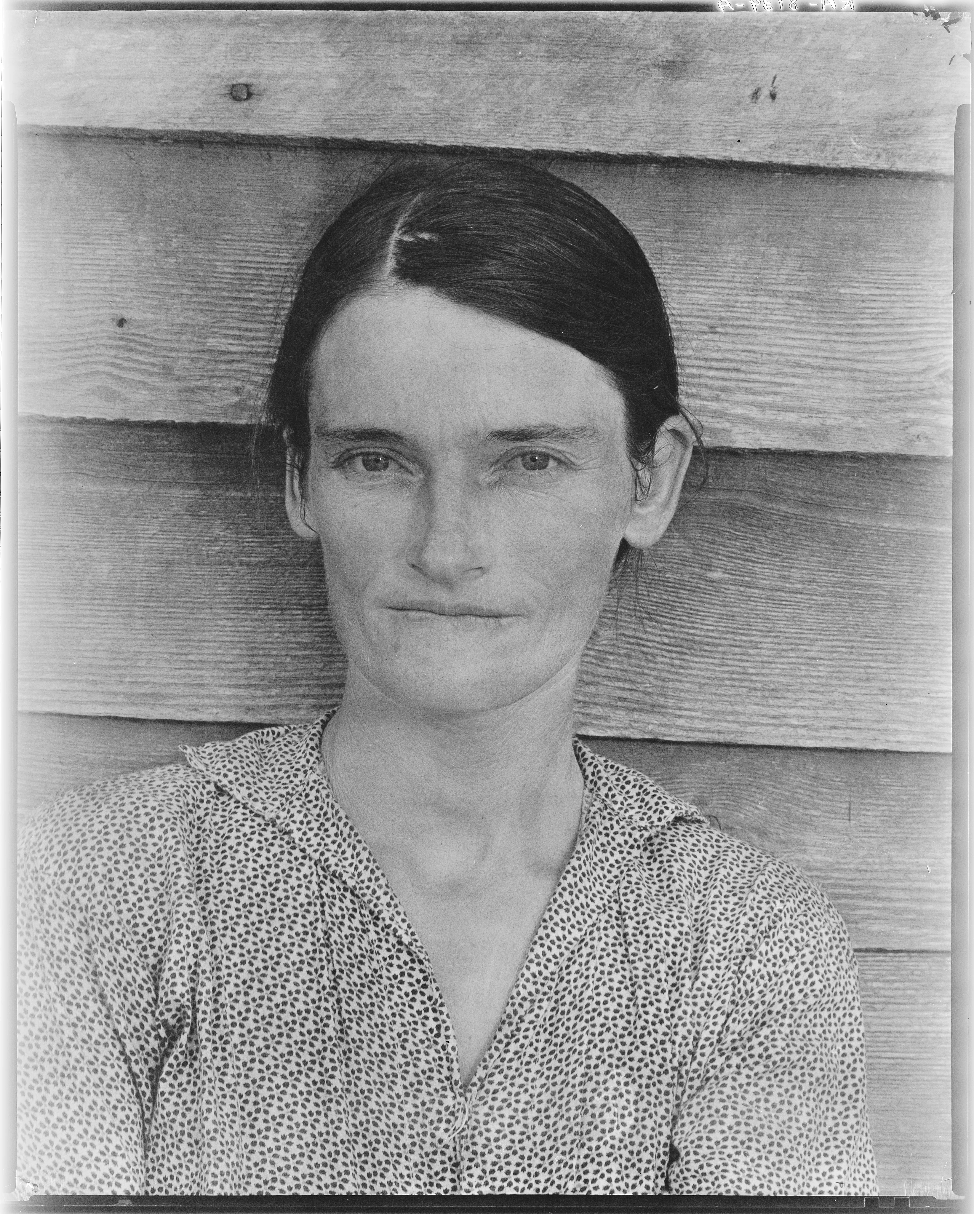 A woman faces the camera directly against a wooden wall.