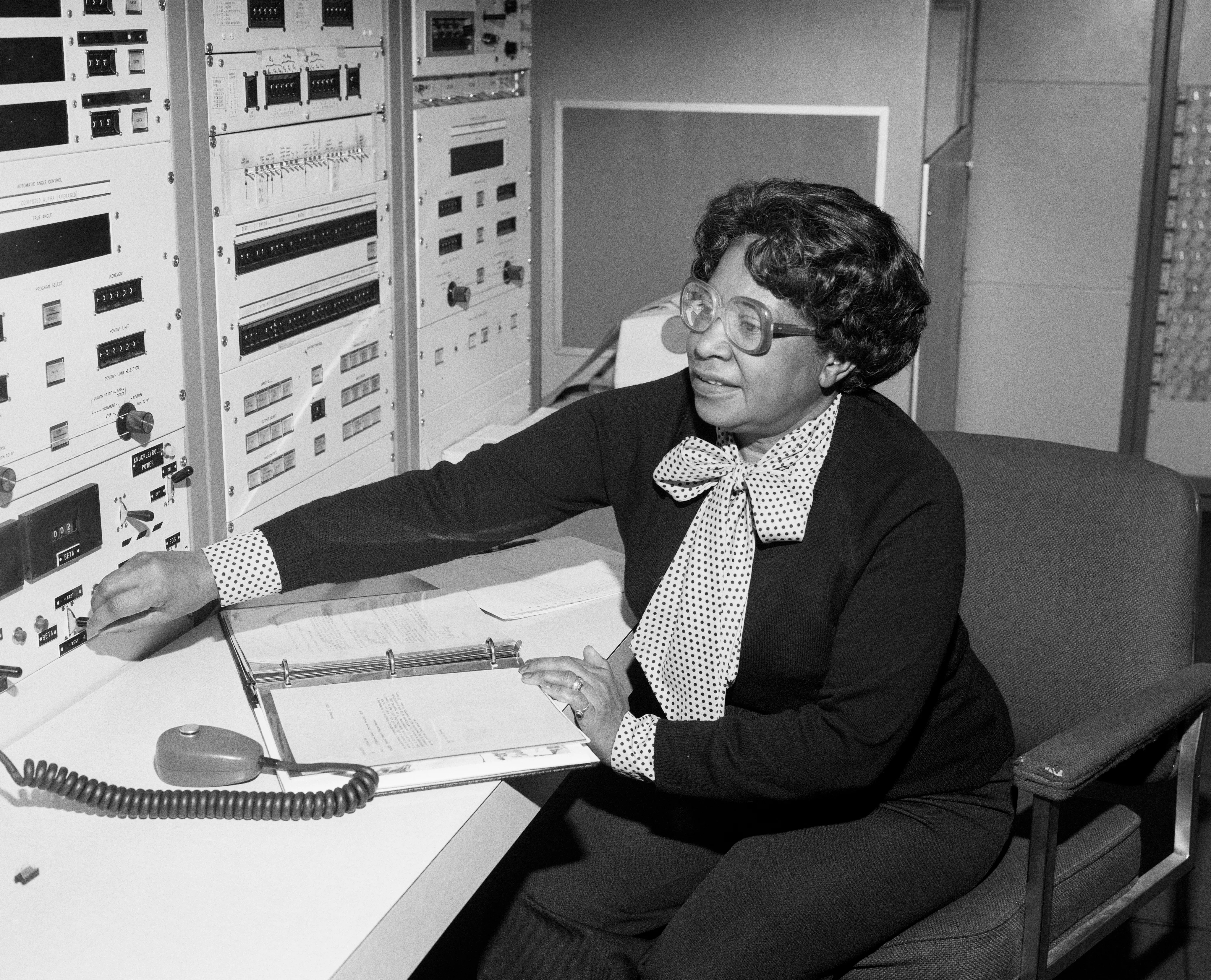 Mary Jackson seated at a desk, reaching toward a tall rack of instruments, with an open binder and a coiled microphone on the table.